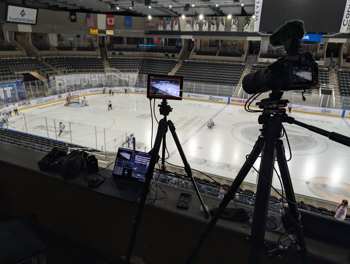 Summer's professional broadcasting setup at the Fargo arena—laptop running OBS Studio, external monitor, and cameras on tripods overlooking the ice